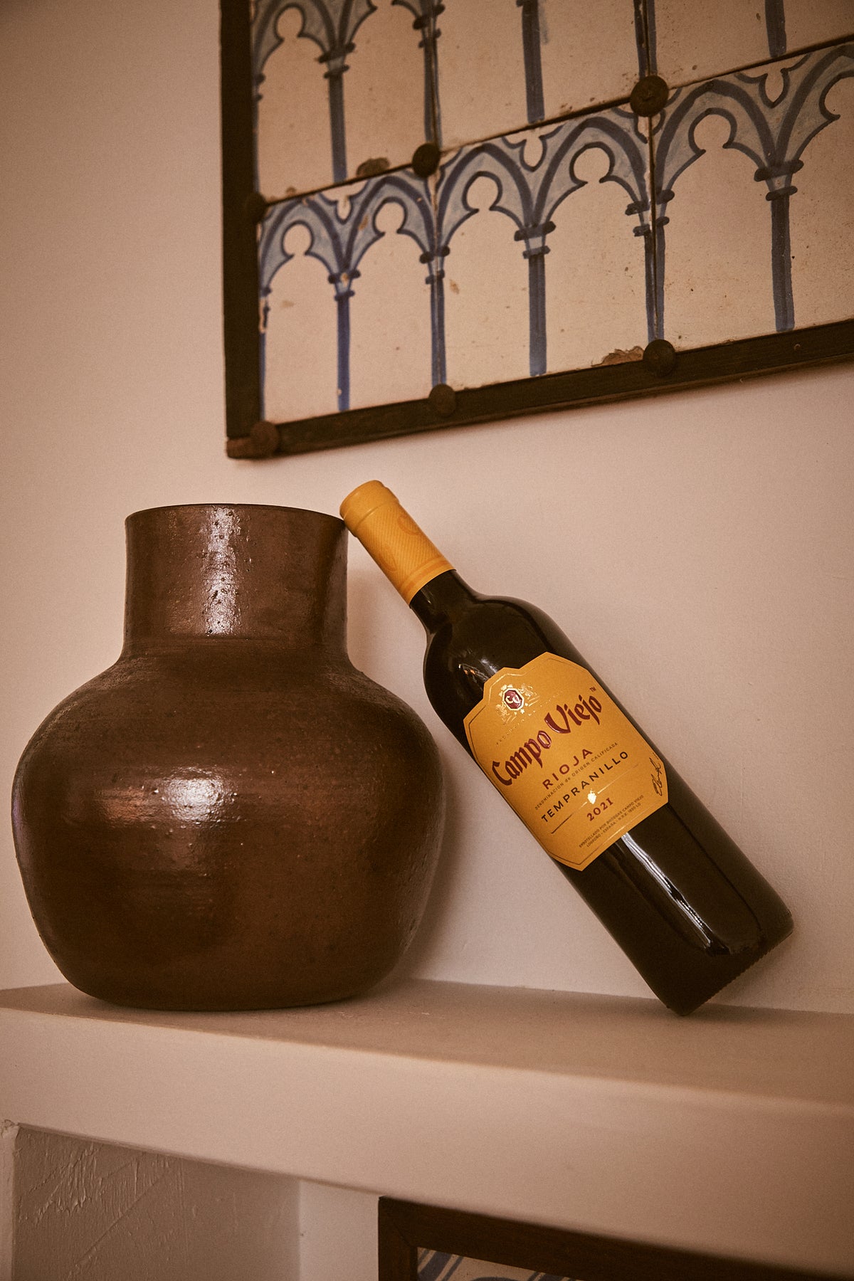 Wine bottle with a yellow label next to a bronze vase on a shelf, with decorative wall art in the background.