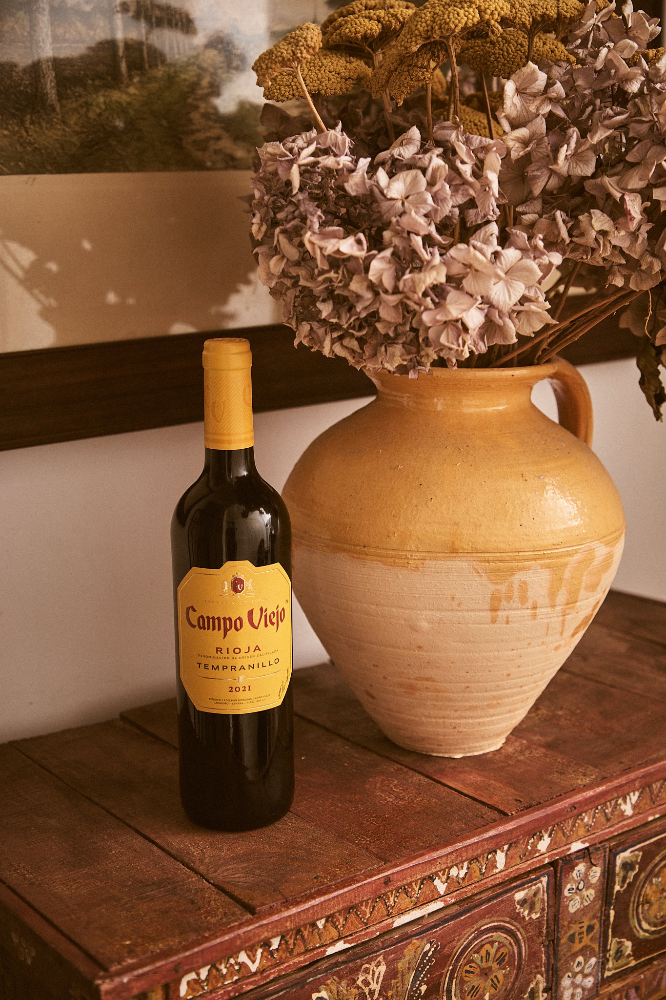 Wine bottle labeled 'Campo Viejo' next to a ceramic vase with dried flowers on a wooden surface.